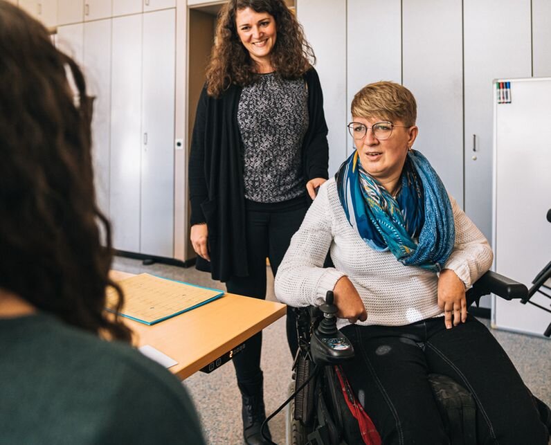 Zwei Frauen. Die Vordere sitzt im Rollstuhl und spricht gerade.