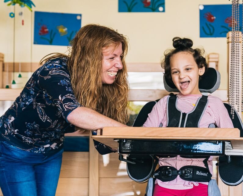 Eine Frau und ein Mädchen lachen gemeinsam. Das Mädchen sitzt im Rollstuhl.