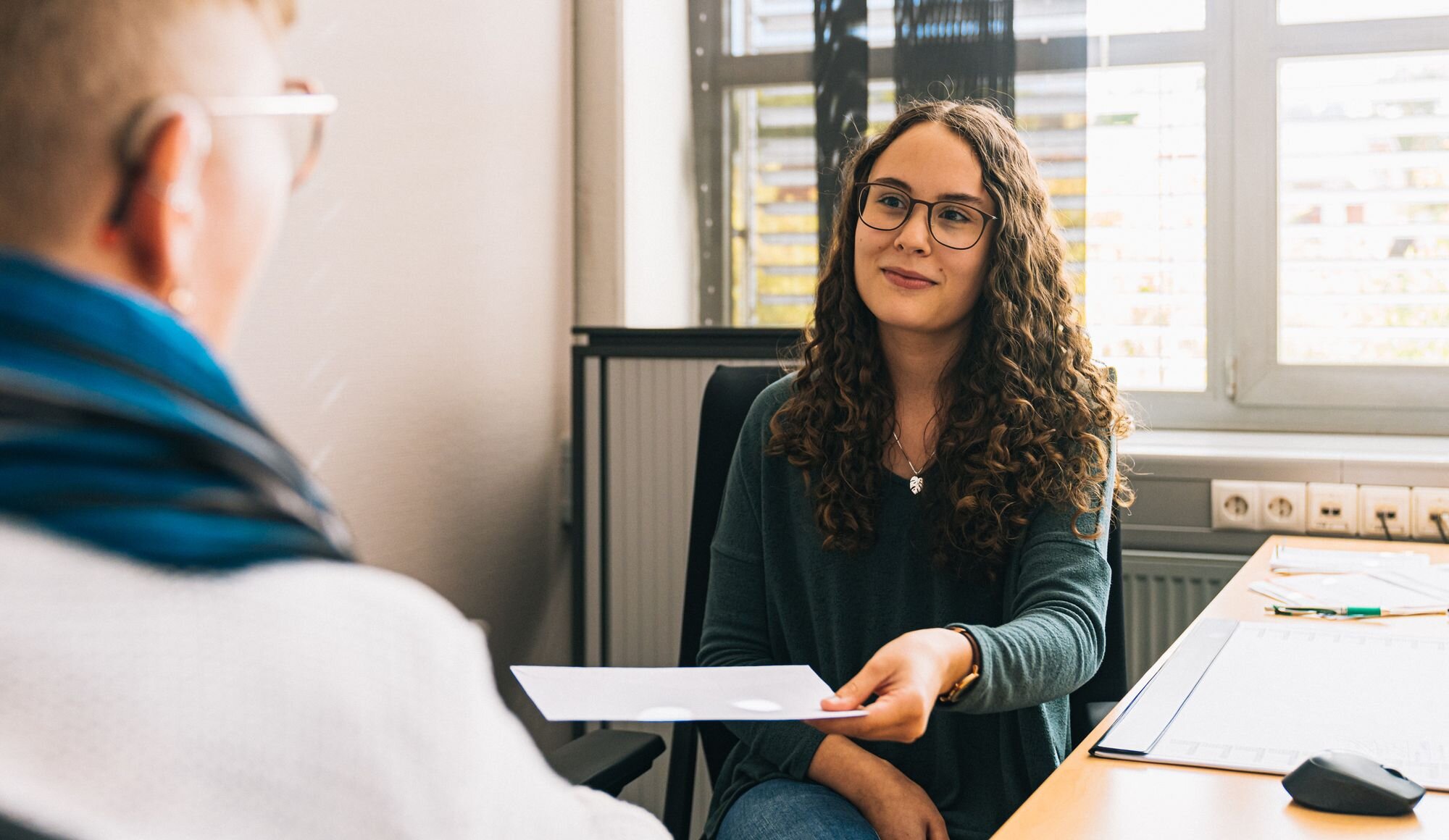 Eine Frau mit Brille übergibt ein Blatt Papier an eine andere Person.